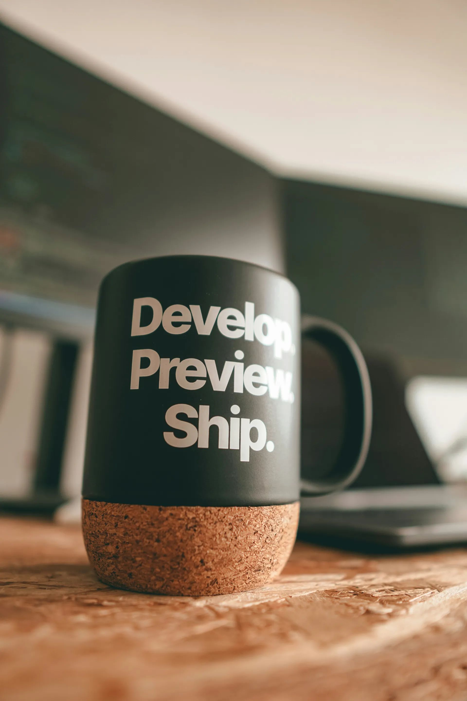 Mok met de tekst 'Develop. Preview. Ship.' naast een laptop op een werkplek