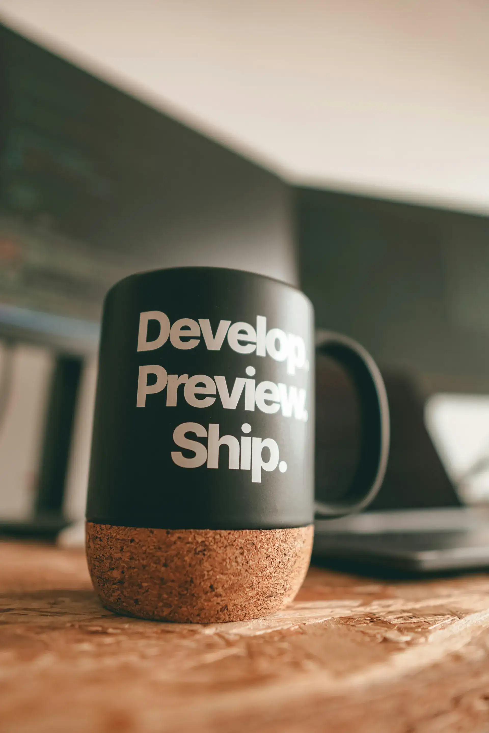 A mug reading 'Develop. Preview. Ship.' beside a laptop on a desk